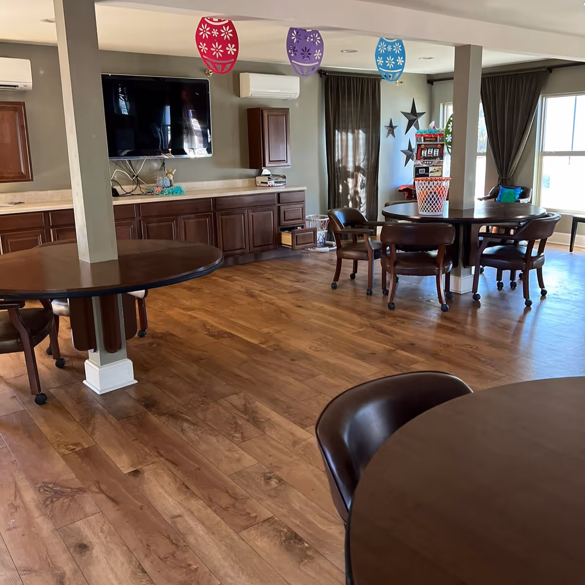 Spacious common room with round tables and chairs, hardwood floors, a wall-mounted TV, and cabinets.