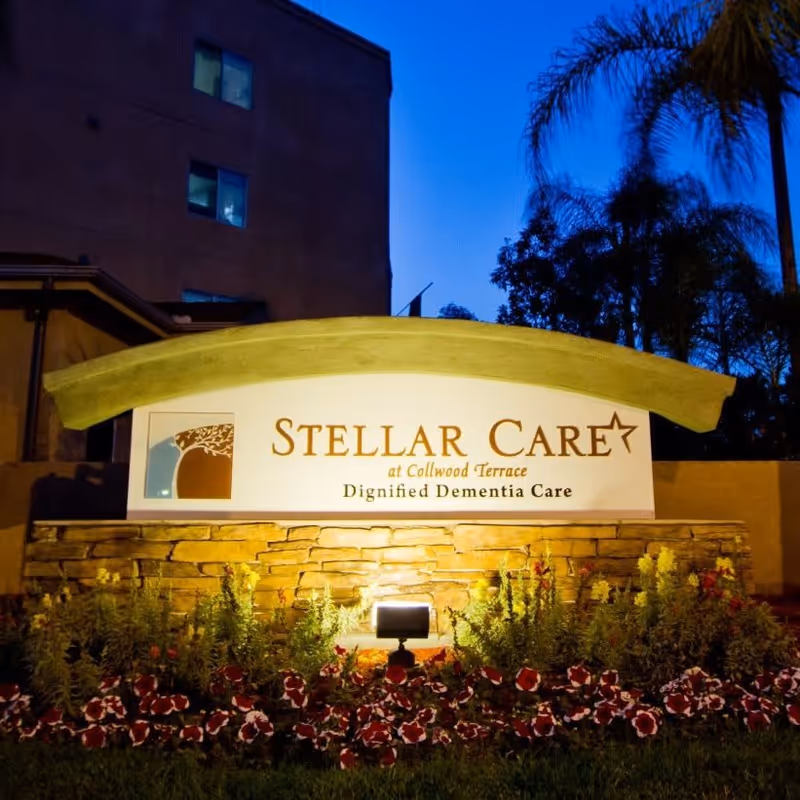 Illuminated outdoor sign for Stellar Care at Collwood Terrace, a facility offering dignified dementia care, surrounded by flowers and plants with a building and trees in the background at dusk.