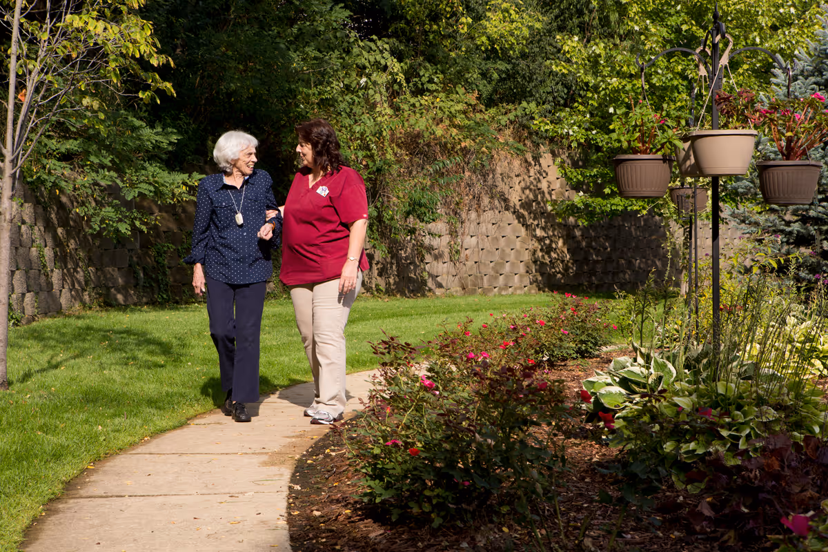 An elderly woman and a caregiver walking arm in arm along a garden path surrounded by green trees, flowering plants, and hanging flower pots on a sunny day.