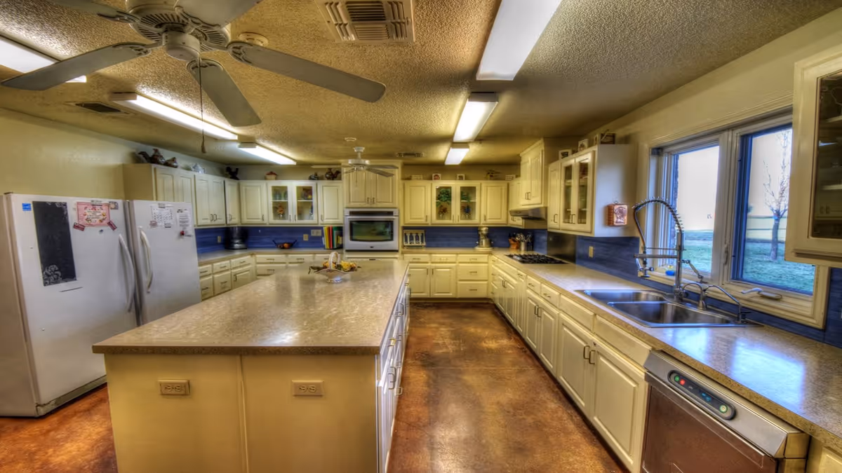 Large bright kitchen with a long center island, white cabinets, double sink by a window, refrigerator and ceiling fans.
