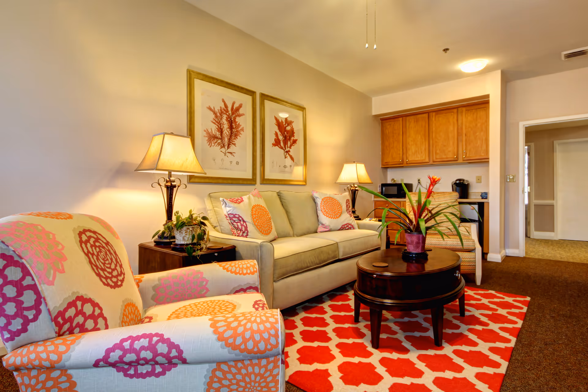 A cozy living room with a beige sofa adorned with colorful floral-patterned pillows, a matching armchair with orange and purple floral designs, two table lamps on either side of the sofa, a round wooden coffee table with a potted plant, and a red patterned rug. The background shows wooden cabinets, a microwave, and a coffee maker, with an open doorway leading to another room.