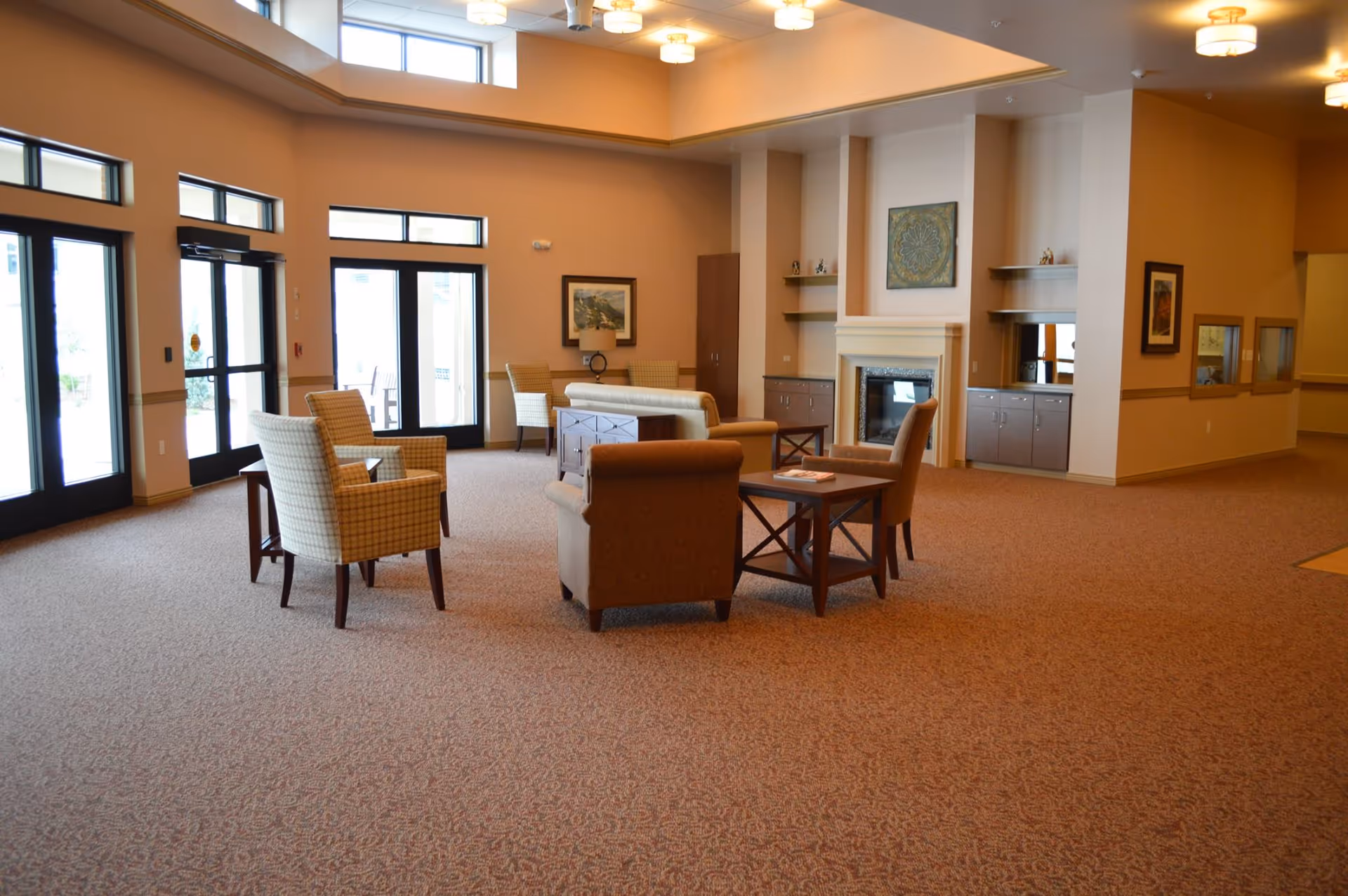 A spacious senior living community lounge area with several armchairs and small tables arranged on a carpeted floor. The room features large windows and glass doors allowing natural light to enter, a fireplace with built-in shelves and cabinets, and warm beige walls with framed artwork.