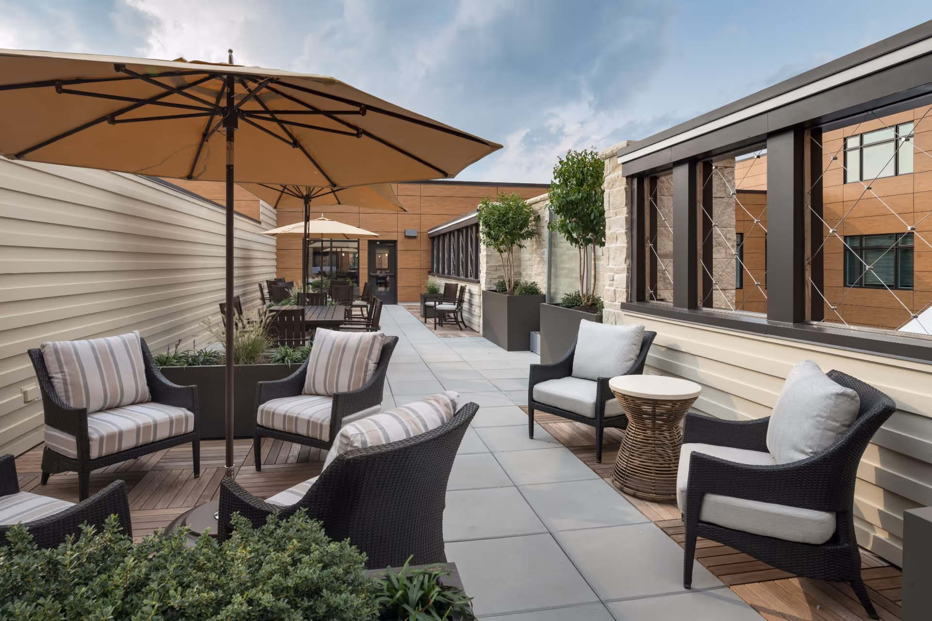 Outdoor patio area with cushioned wicker chairs, small tables, large beige umbrellas, potted plants, and a tiled floor, adjacent to a building with beige siding and stone accents under a partly cloudy sky.