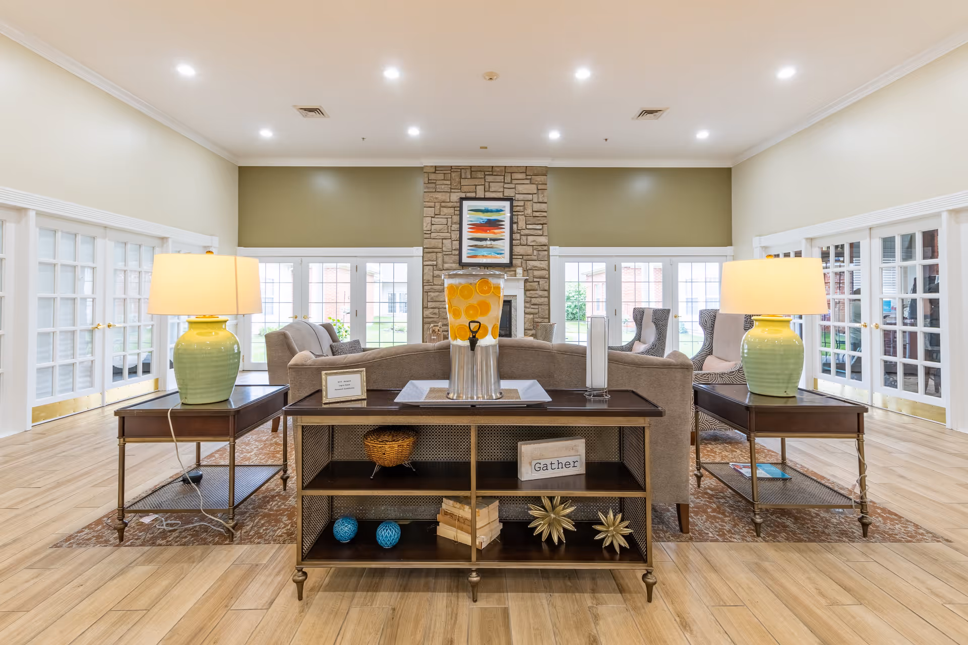A bright and spacious living room area in Walnut Ridge Assisted Living and Memory Care featuring a stone fireplace with a colorful abstract painting above it. The room has large windows and glass doors letting in natural light. There are two beige sofas and two patterned armchairs arranged around the fireplace. Two side tables with green ceramic lamps flank the sofas. A wooden console table with decorative items including a basket, books, and a sign that says 'Gather' is positioned behind one sofa. The floor is light wood and the walls are painted in neutral tones.
