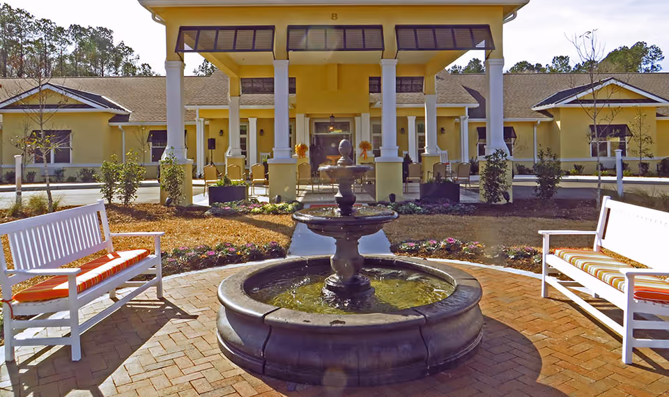 Outdoor view of The Addison of Bluffton facility entrance featuring a circular water fountain in the foreground, two white benches with orange and white striped cushions on either side, and a yellow building with a covered porch supported by white columns in the background.