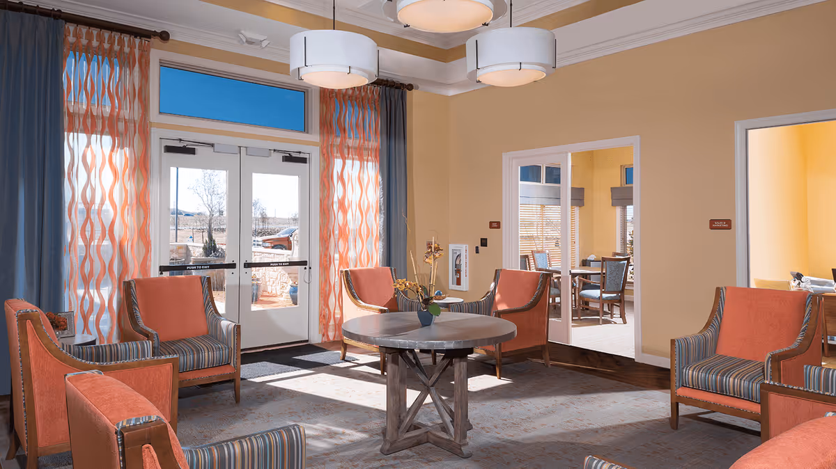 A bright and inviting common area with several orange and striped armchairs arranged around a round wooden table with a small flower arrangement. The room has large windows and glass doors with orange and blue curtains, allowing natural light to fill the space. Adjacent rooms with dining tables and chairs are visible through open doorways.