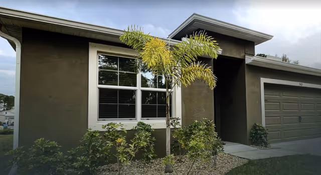 Front exterior of a single-story building with a large window, small palm tree, shrubs, and an attached garage.