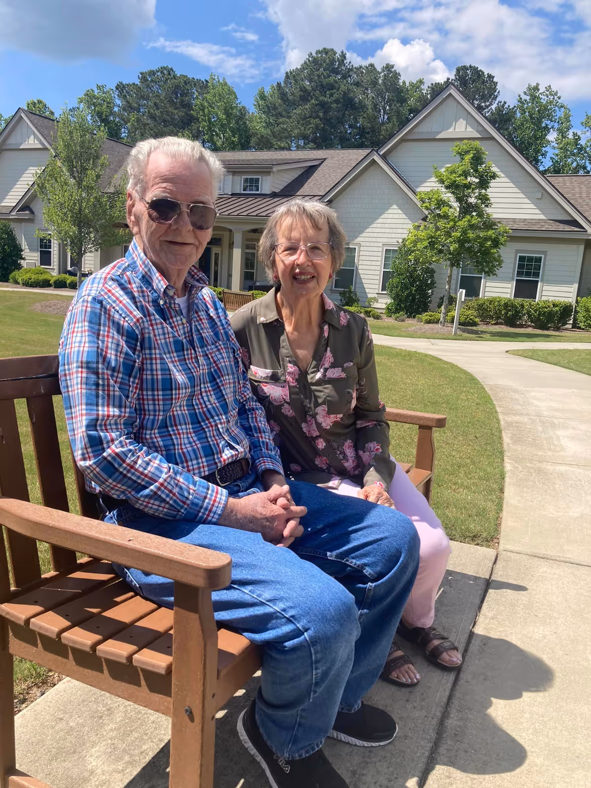 An elderly man and woman sitting on a wooden bench outside on a sunny day, with a senior living community building and green trees in the background. The man is wearing sunglasses, a plaid shirt, and jeans, while the woman is wearing glasses, a floral blouse, and light-colored pants.