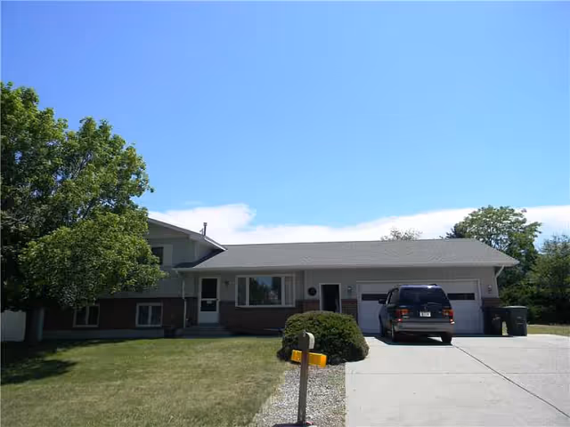 Front exterior view of a single-story residential building with a two-car garage, a driveway with a parked black SUV, a green lawn, a tree on the left side, and a clear blue sky.