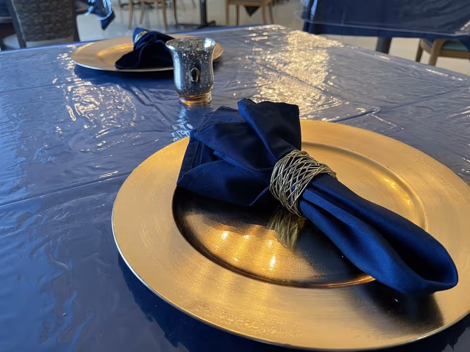 Close-up of a dining table set with a gold charger plate and a navy blue cloth napkin held by a silver napkin ring. The table is covered with a shiny blue tablecloth, and another similarly set place is visible in the background along with chairs and part of the dining area.