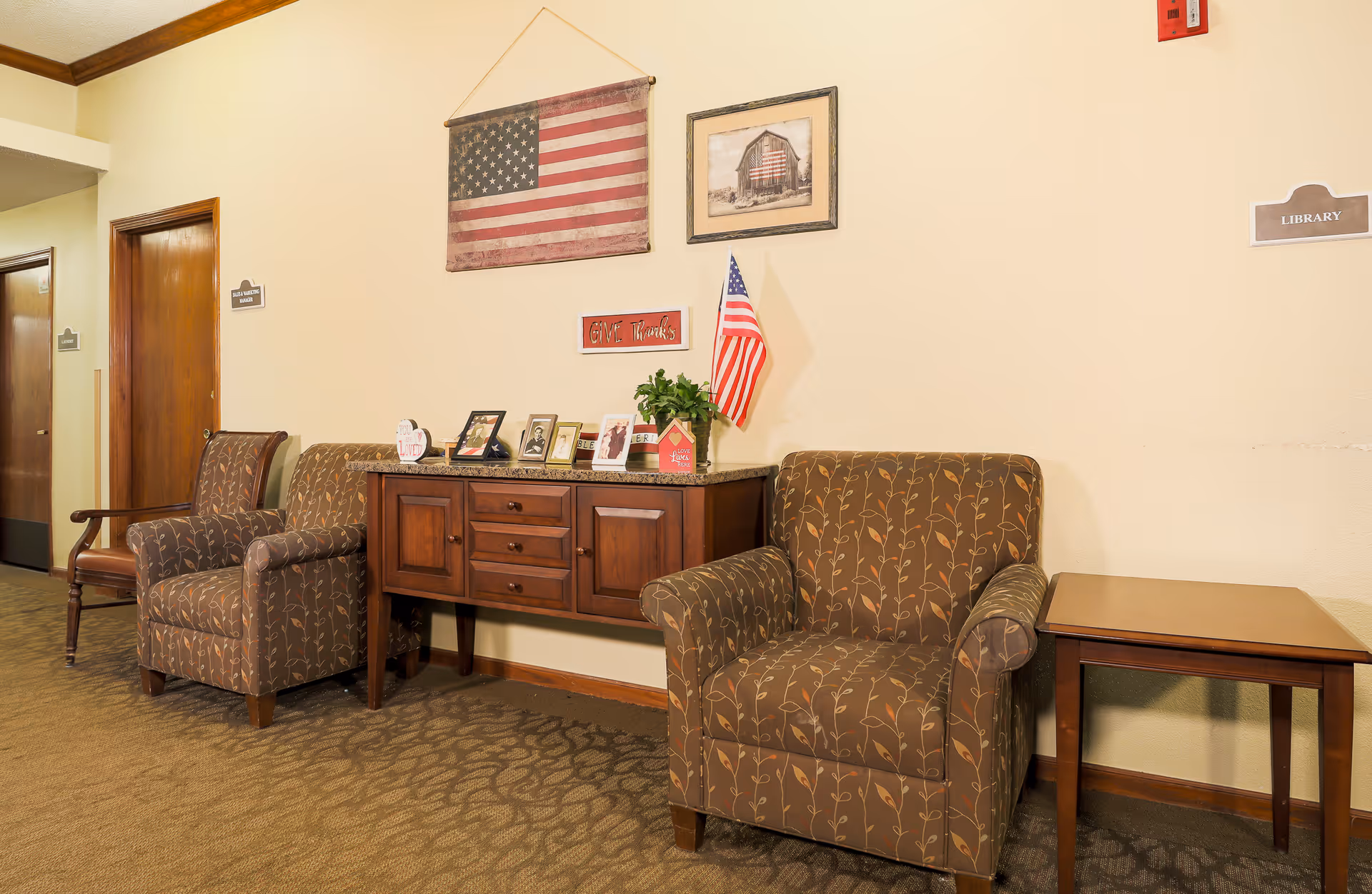 A cozy seating area in a hallway with two patterned armchairs and a wooden side table. Between the chairs is a wooden cabinet displaying framed photos, a small plant, and an American flag. Above the cabinet hangs a large American flag and a framed picture of a barn with an American flag painted on it. The walls are light-colored, and there are signs indicating directions to rooms such as the library.