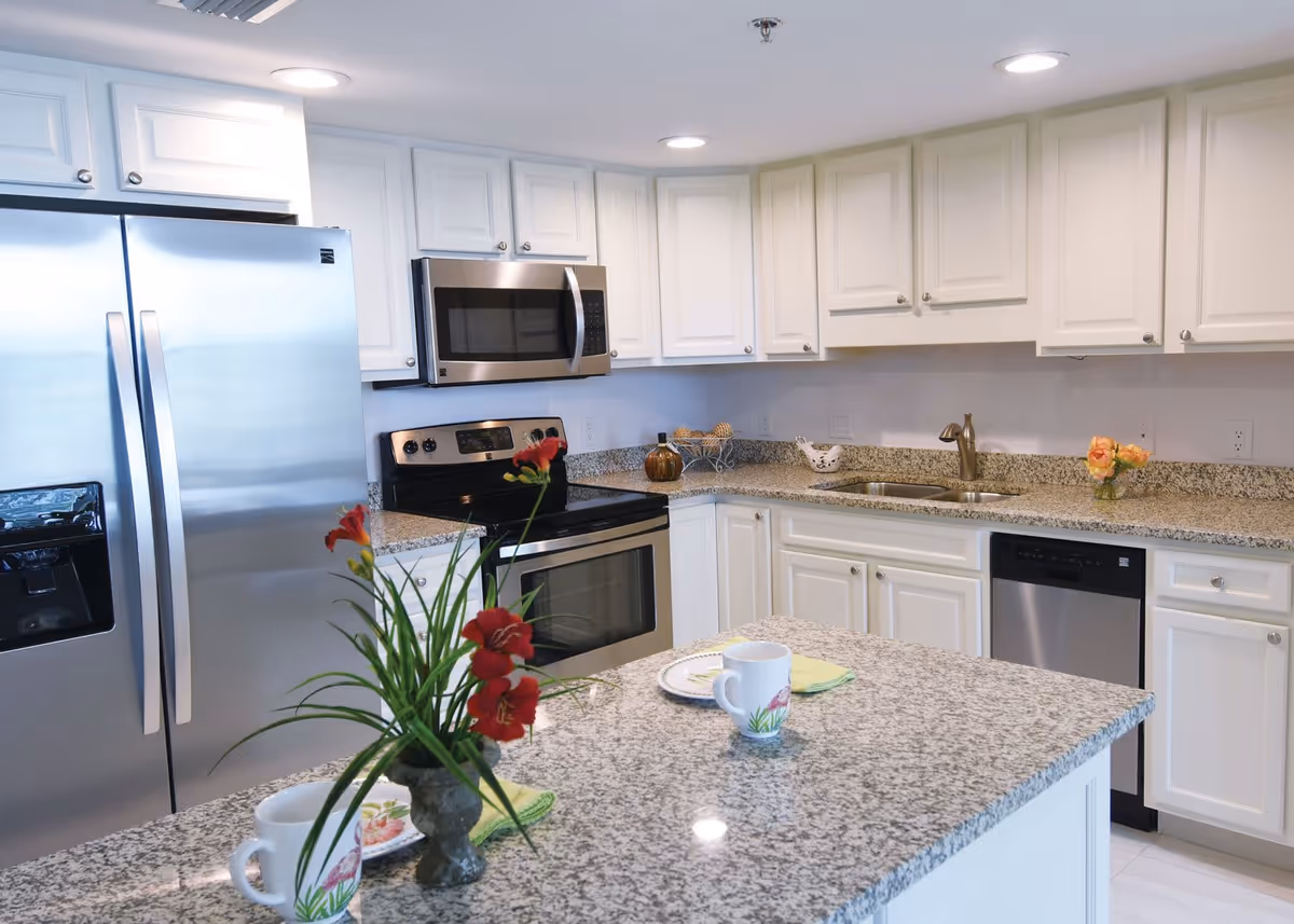 A modern kitchen with white cabinets and granite countertops. The kitchen features stainless steel appliances including a refrigerator, microwave, oven, and dishwasher. There is a kitchen island with a vase of red flowers, two coffee mugs, and plates with green napkins. The space is well-lit with recessed ceiling lights.