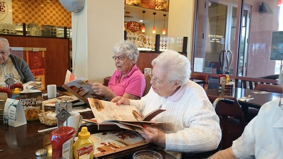 Three elderly individuals sitting at a dining table in a restaurant or dining area, looking at menus. The table has condiments including ketchup and mustard, a coffee cup, and a sign advertising Dutch apple pie. The background shows a counter area with warm lighting and a glass door entrance.