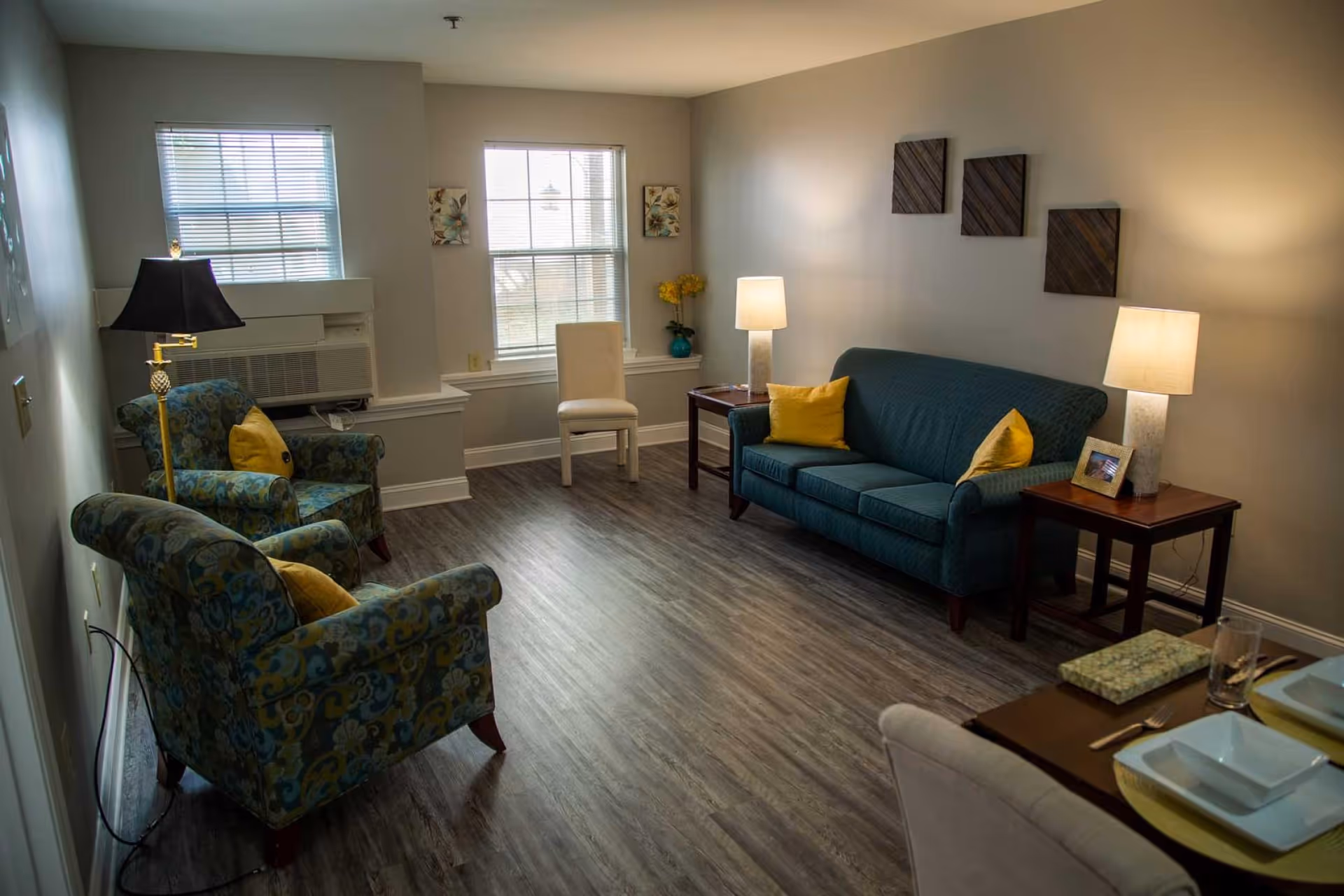 A cozy living room with two patterned armchairs and a blue sofa, each with yellow accent pillows. There are two windows with blinds, two side tables with lamps, a white chair near the window, and decorative wall art. The floor is wood, and a small dining table with place settings is partially visible in the foreground.