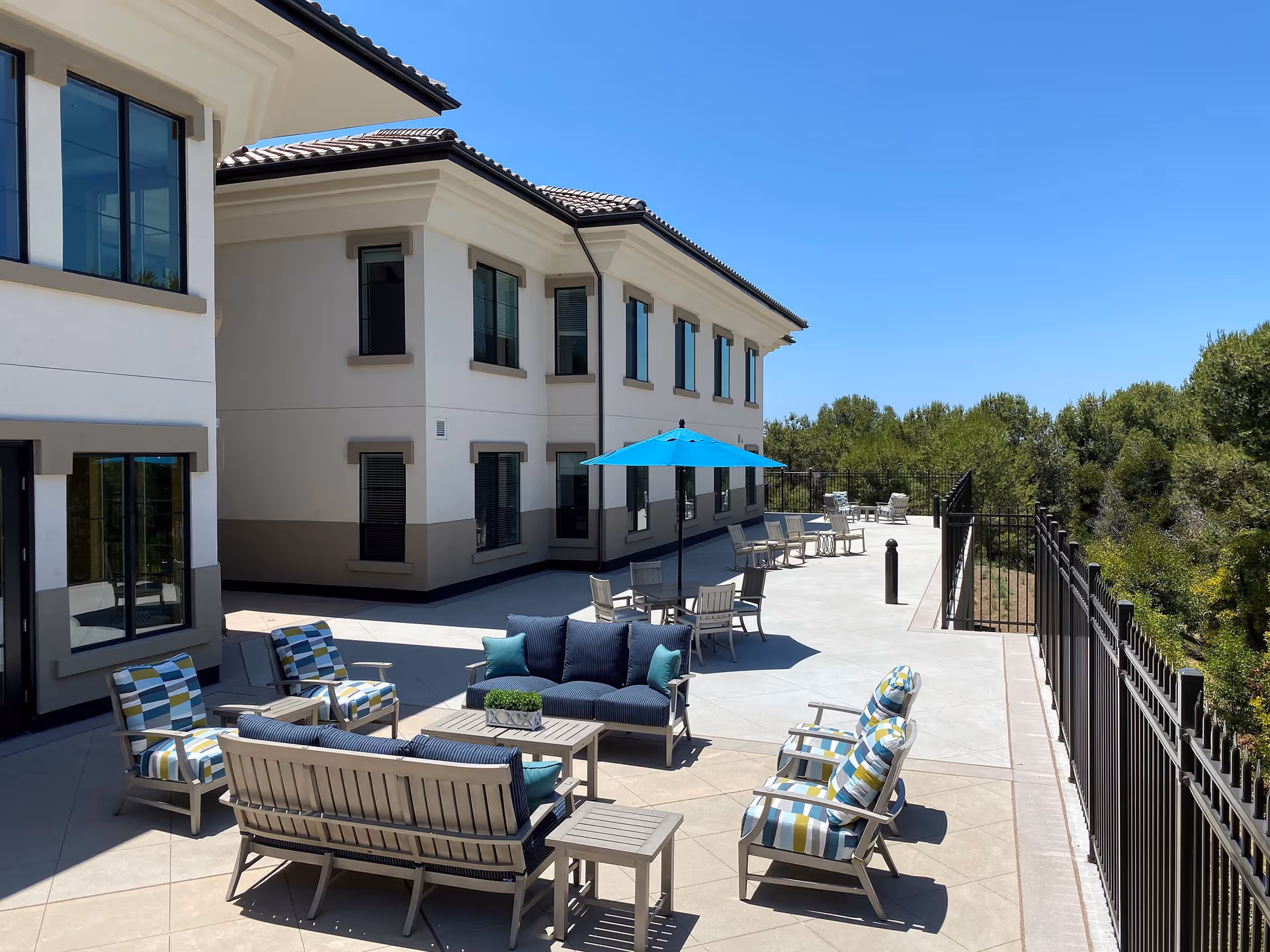 Outdoor patio area at Silvergate Rancho Bernardo featuring multiple seating arrangements with cushioned chairs and sofas, a table with a blue umbrella, and a black metal fence along the edge overlooking greenery under a clear blue sky.