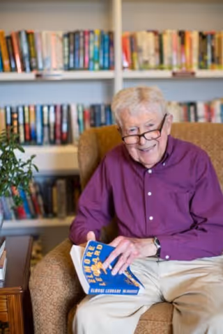 An elderly man wearing glasses and a purple shirt sits in a comfortable armchair in front of a bookshelf filled with books. He is smiling and holding a blue book titled 'Elmore Leonard'.