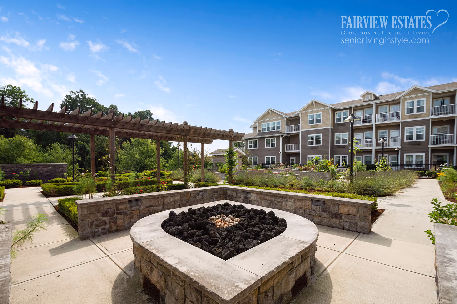 Courtyard with a stone fire pit, pergola, landscaped walkways, and a multi-story senior living building under a blue sky.