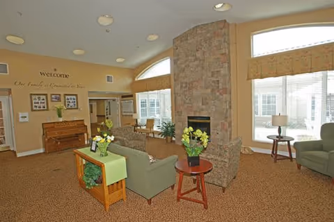 Community living room with sofas and chairs arranged around a stone fireplace, a piano against the wall, large windows, and decorative plants.