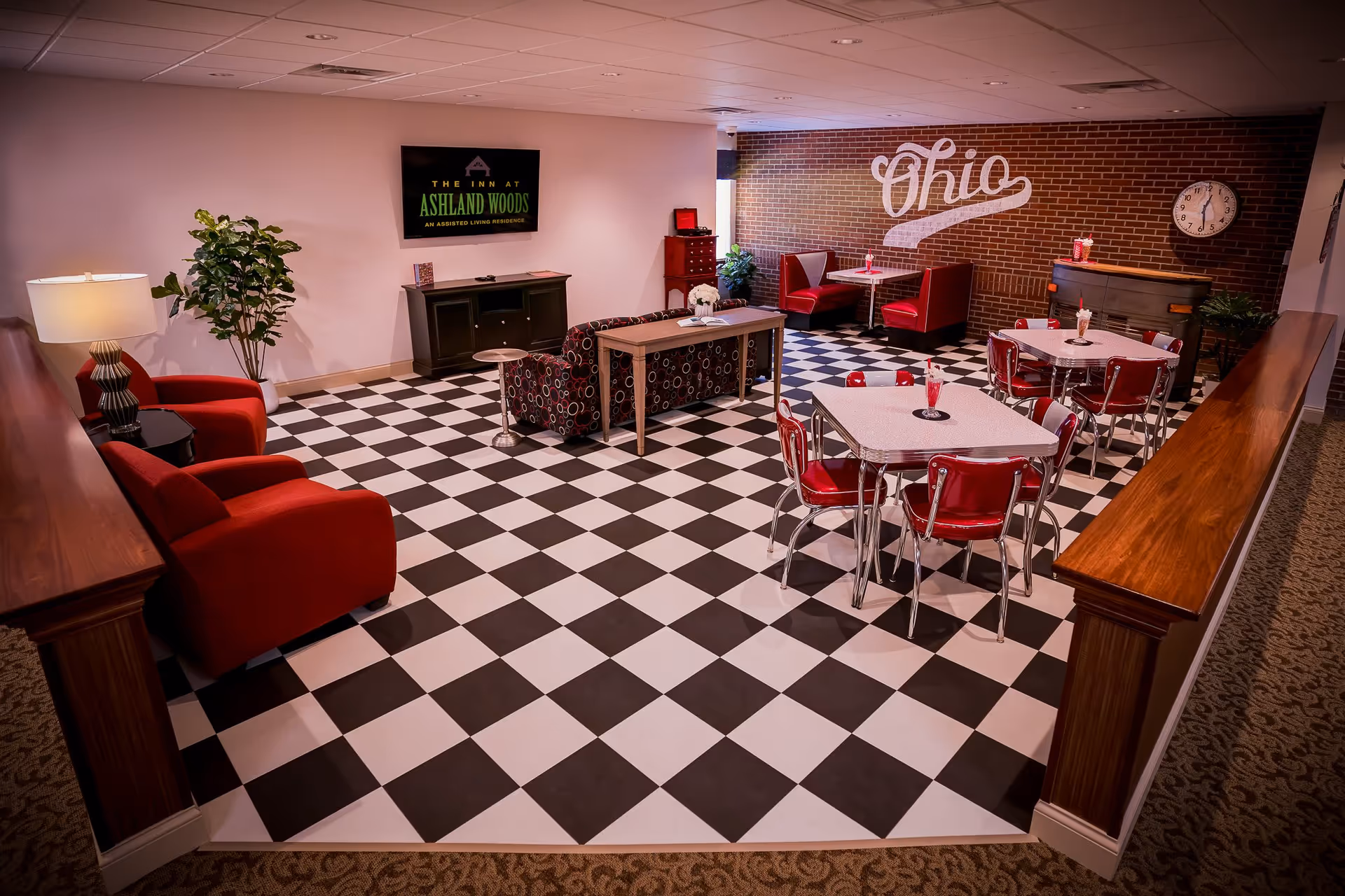 A retro-style common area with black and white checkered flooring, red chairs and booths, tables with milkshakes, a patterned sofa, a TV mounted on the wall displaying 'The Inn at Ashland Woods', a brick wall with the word 'Ohio' painted on it, a clock, and some plants.
