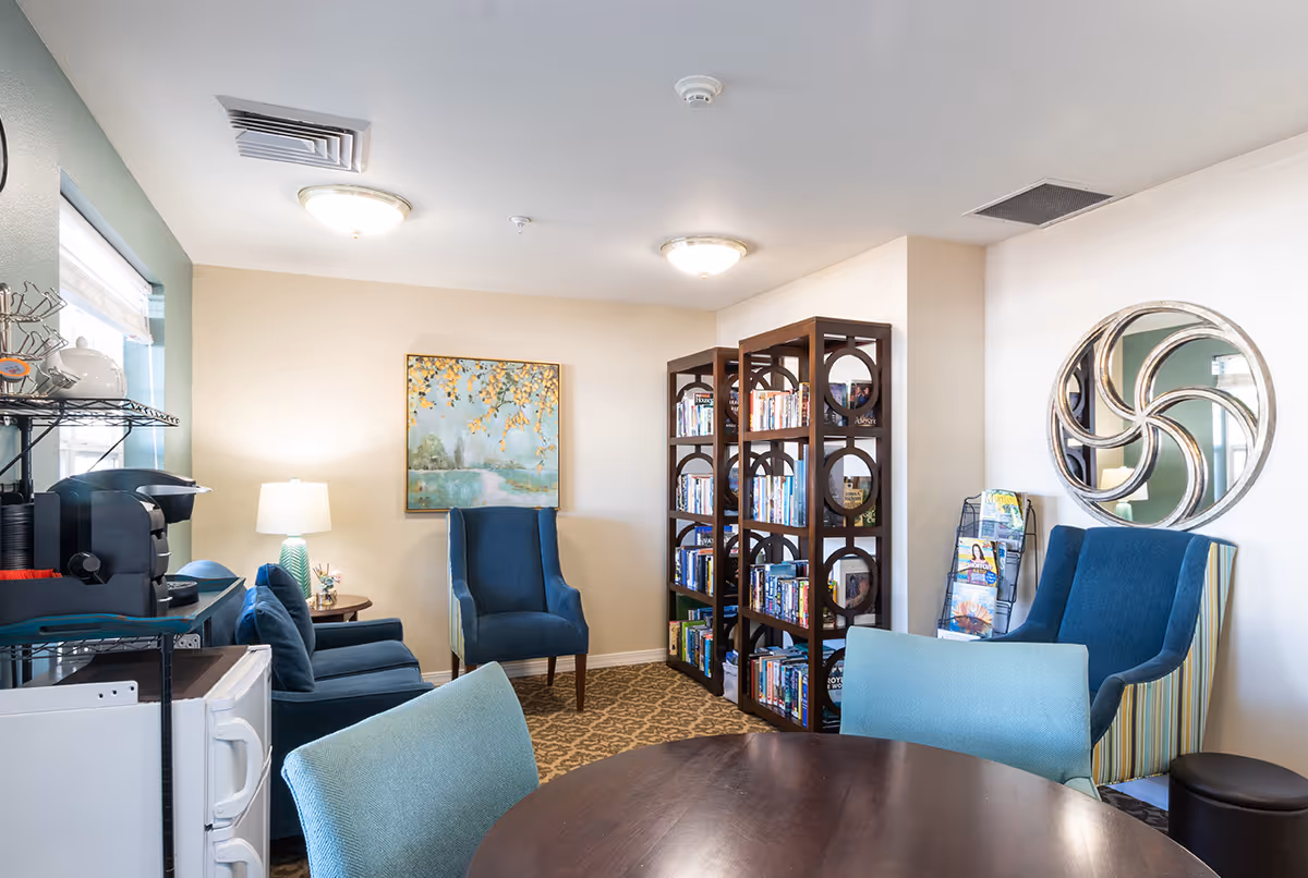 A cozy senior living common area with blue upholstered chairs, a round wooden table with light blue chairs, a bookshelf filled with books, a coffee station with a coffee maker and cups, a lamp on a side table, a decorative wall mirror, and a framed painting on the wall.
