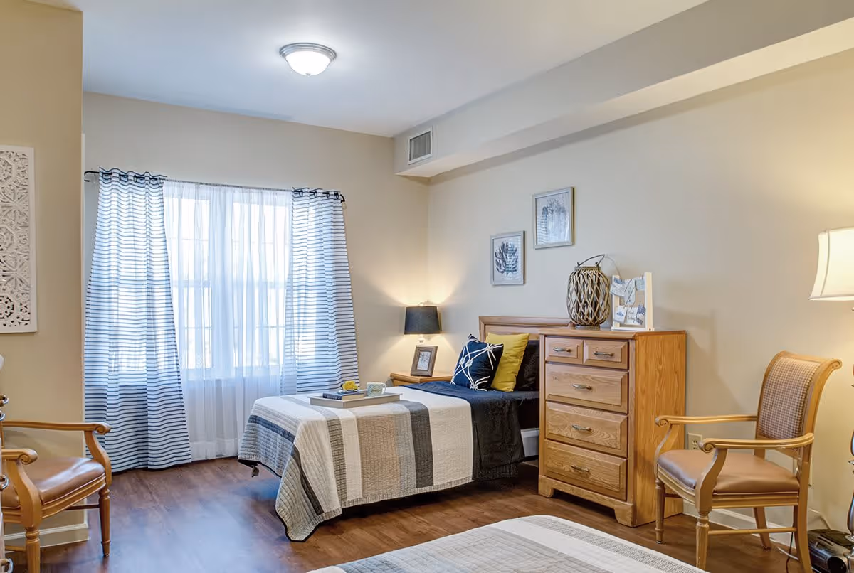 A well-lit bedroom with two single beds covered in striped quilts. The room features wooden furniture including a dresser and two chairs with armrests. There are two framed botanical prints on the wall above the dresser, a table lamp on a nightstand, and a window with sheer and striped curtains allowing natural light to enter.