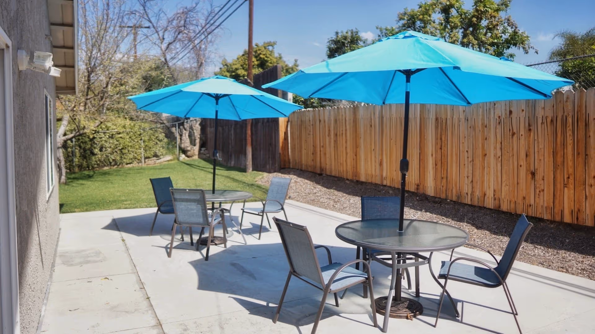 Outdoor patio area with two round glass tables, each shaded by a large blue umbrella. Each table is surrounded by four metal chairs with mesh seats and backs. The patio is adjacent to a building with a beige exterior wall and is bordered by a wooden fence and some greenery in the background under a clear blue sky.