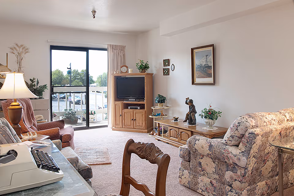 A cozy living room with floral patterned armchairs, a wooden TV cabinet with a television, a wooden side table with decorative items, a lamp on a side table, and a sliding glass door leading to a balcony with a view of trees and parked cars outside.