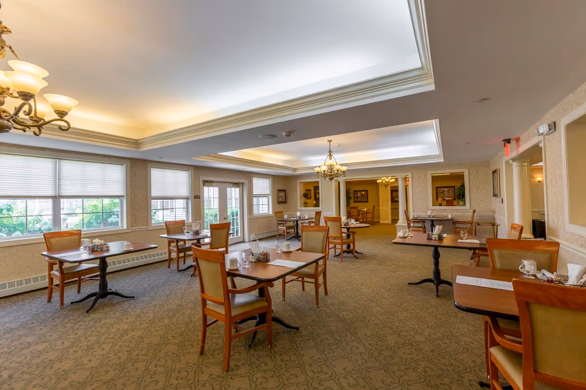 A spacious dining room with multiple wooden tables and chairs arranged neatly. Each table is set with cups, glasses, and napkins. The room has large windows with blinds allowing natural light to brighten the space. Elegant chandeliers hang from the ceiling, and the walls are decorated with framed artwork and mirrors.