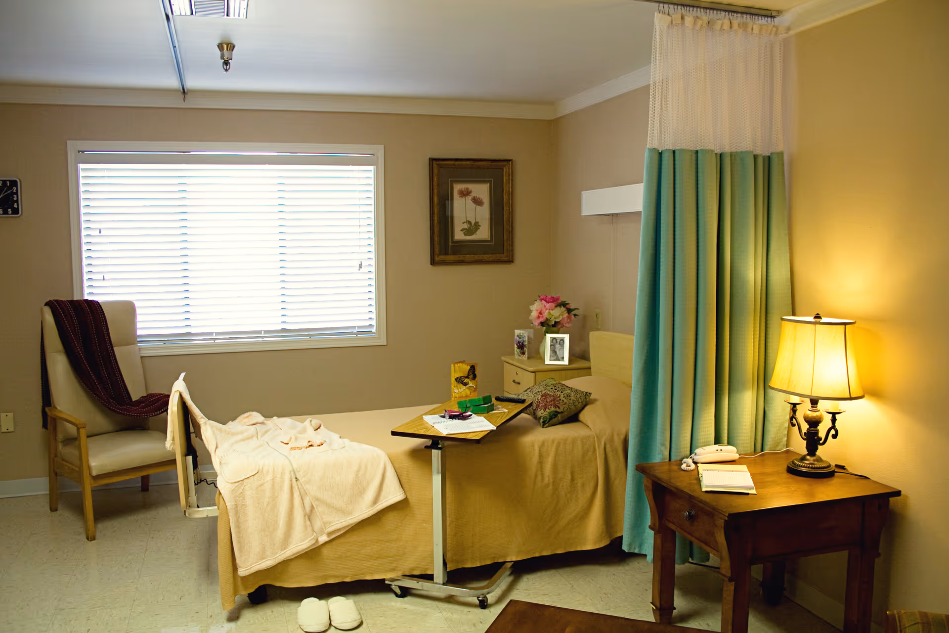 A cozy room with a single bed covered in beige bedding, a small wooden side table with a lamp and a telephone, a chair with a blanket draped over it, a window with blinds, and a curtain partially drawn around the bed for privacy. There are framed pictures and flowers on a small cabinet next to the bed.