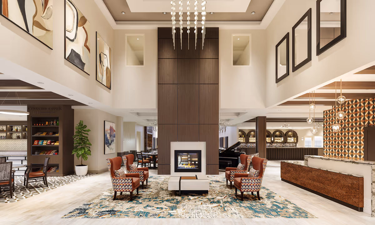 A spacious and modern senior living facility common area with a central fireplace surrounded by four patterned armchairs on a decorative rug. The room features high ceilings with hanging light fixtures, abstract artwork on the walls, a small snack counter area to the left, and a bar with pendant lights and a geometric patterned wall to the right.
