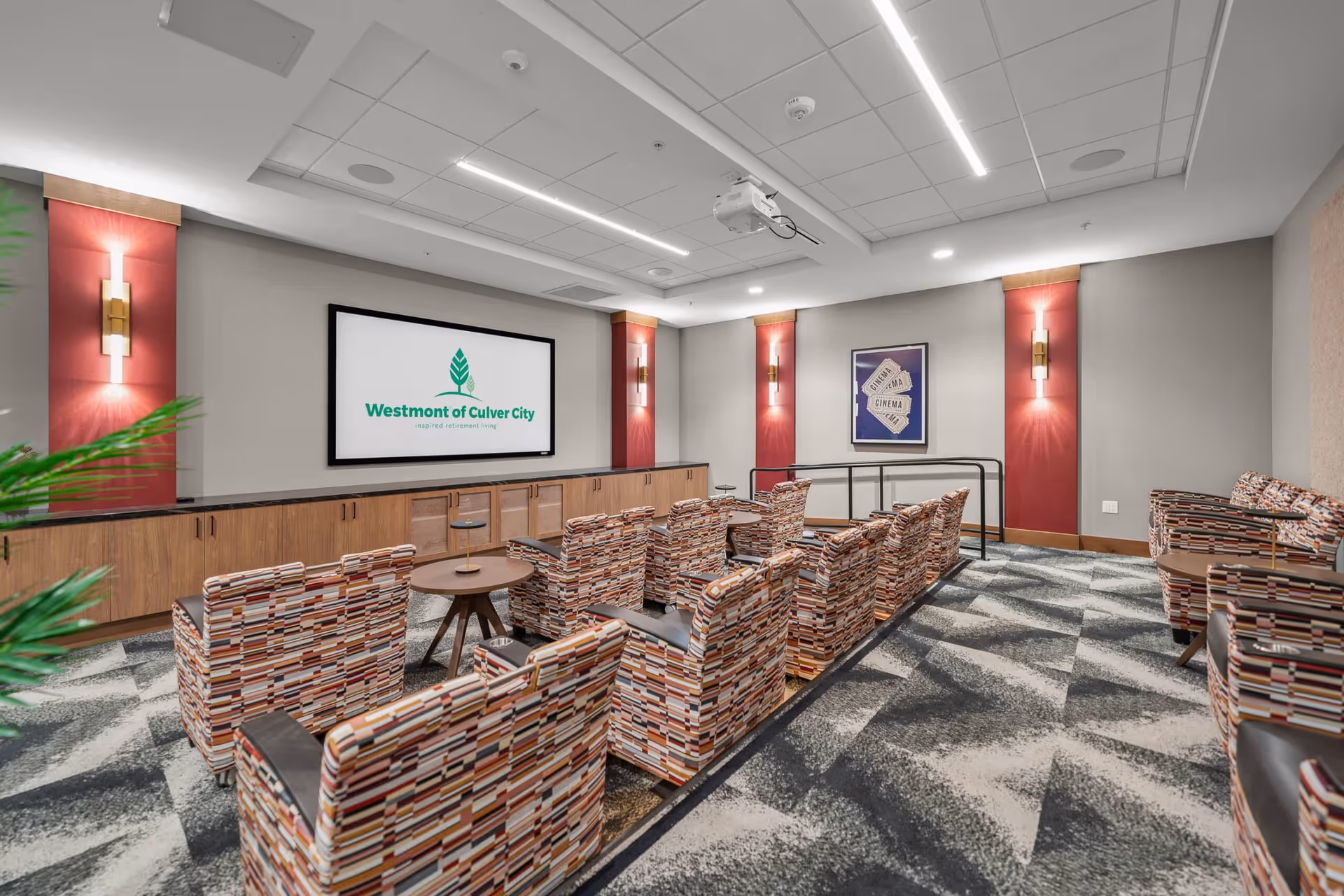 A modern theater room with patterned armchairs arranged in rows facing a large screen displaying the Westmont of Culver City logo. The room features red accent panels with wall sconces, a patterned carpet, and a ceiling-mounted projector.