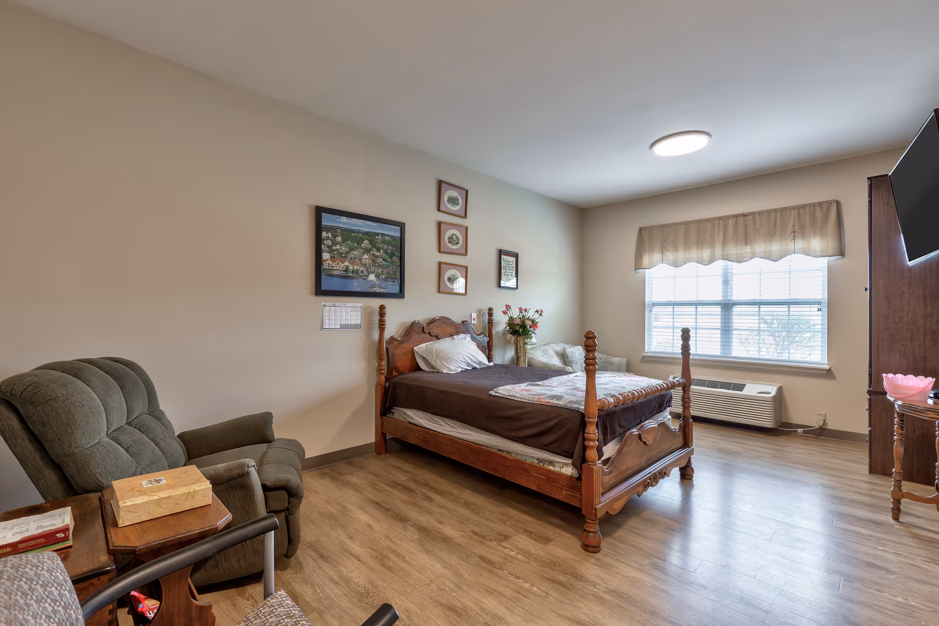 A cozy bedroom with a wooden four-poster bed made with brown bedding, a green recliner chair, a small side table with books and a tissue box, a window with blinds and a valance, a wall-mounted TV, and framed pictures on the wall.
