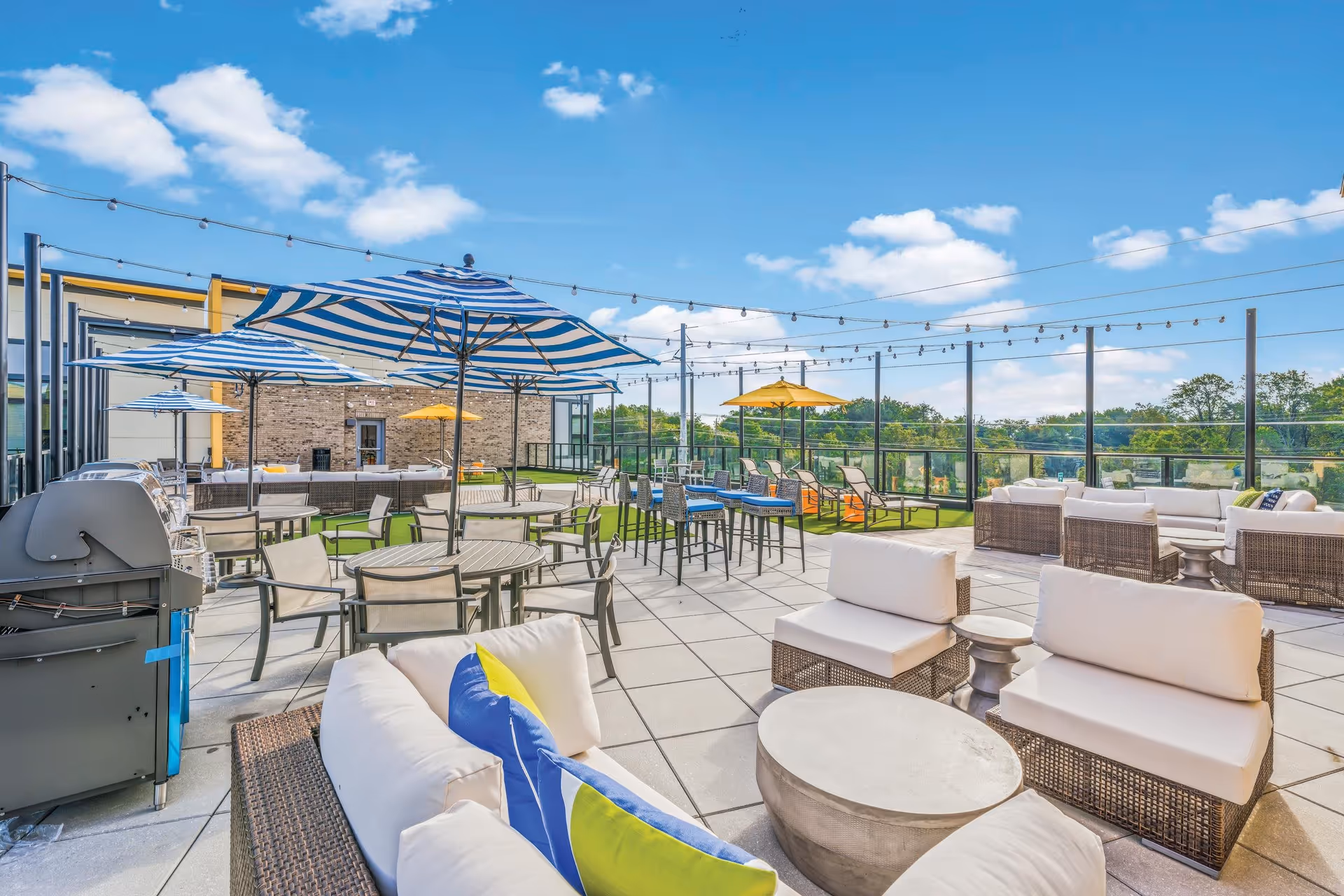 Outdoor patio area with multiple seating arrangements including cushioned wicker chairs, round tables with striped blue and white umbrellas, and lounge chairs under yellow umbrellas. String lights are hung above, and there is a grill on the left side. The area overlooks greenery under a blue sky with scattered clouds.