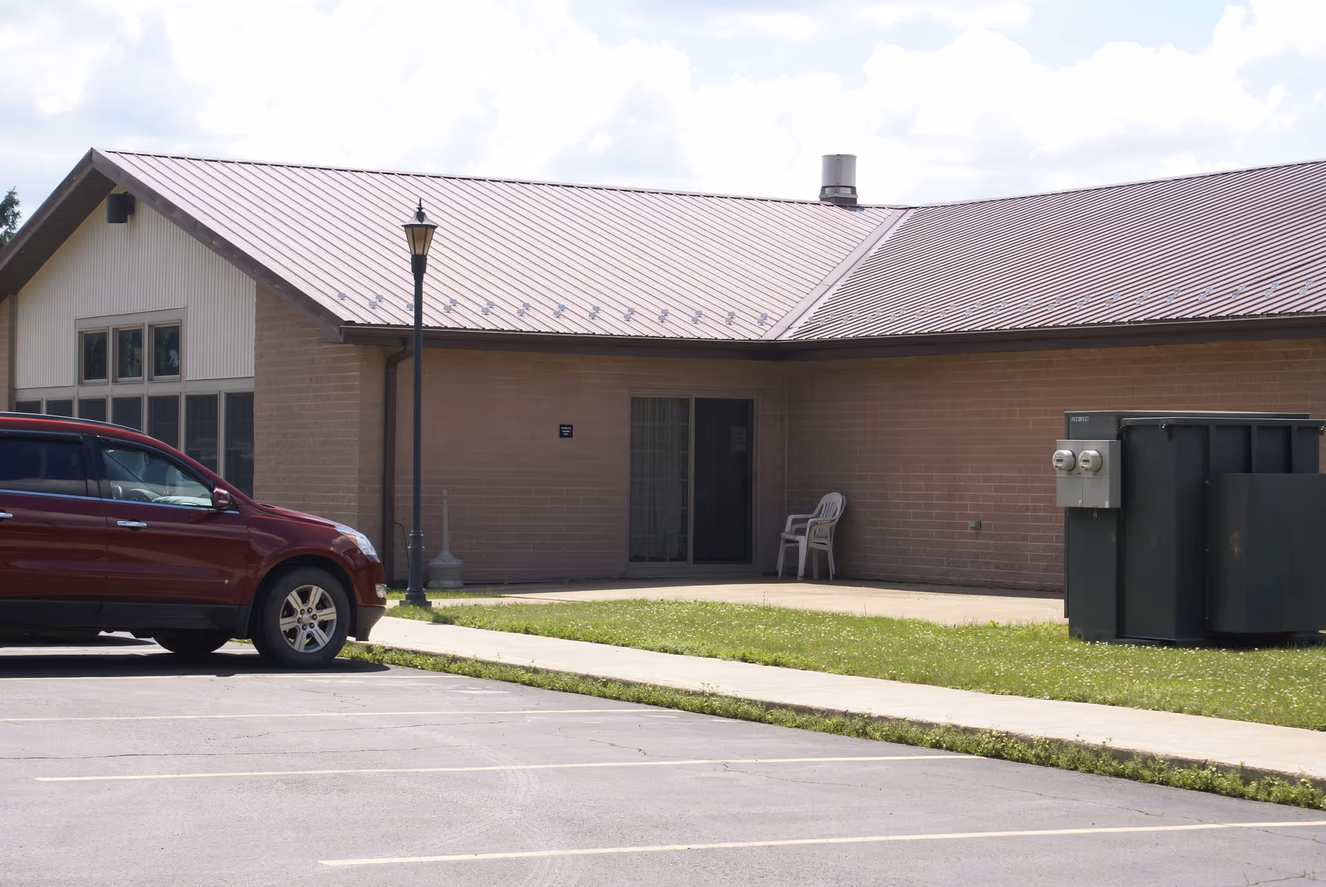 Single-story brick building with a metal roof, a parked red SUV in front, white plastic chairs by a glass door, and an electrical transformer near the parking lot.