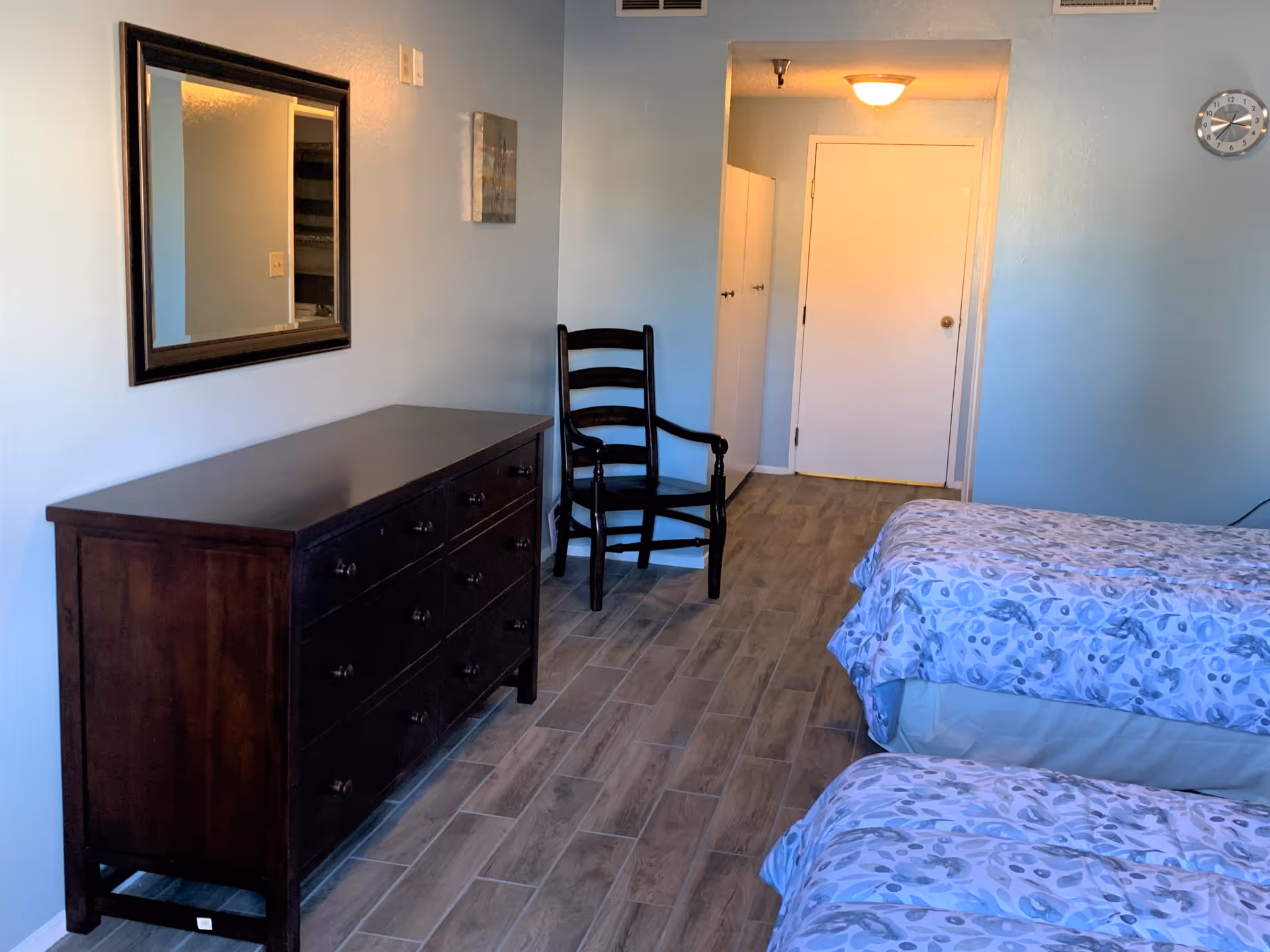 Two beds with blue floral bedding, a dark dresser, and a chair in a blue bedroom.