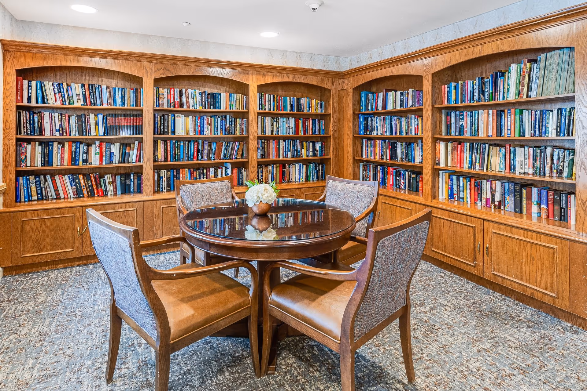 A cozy library room with wooden bookshelves filled with books lining the walls. In the center, there is a round wooden table with a small floral arrangement on top, surrounded by four cushioned chairs with wooden frames. The room has a carpeted floor and a bright, inviting atmosphere.