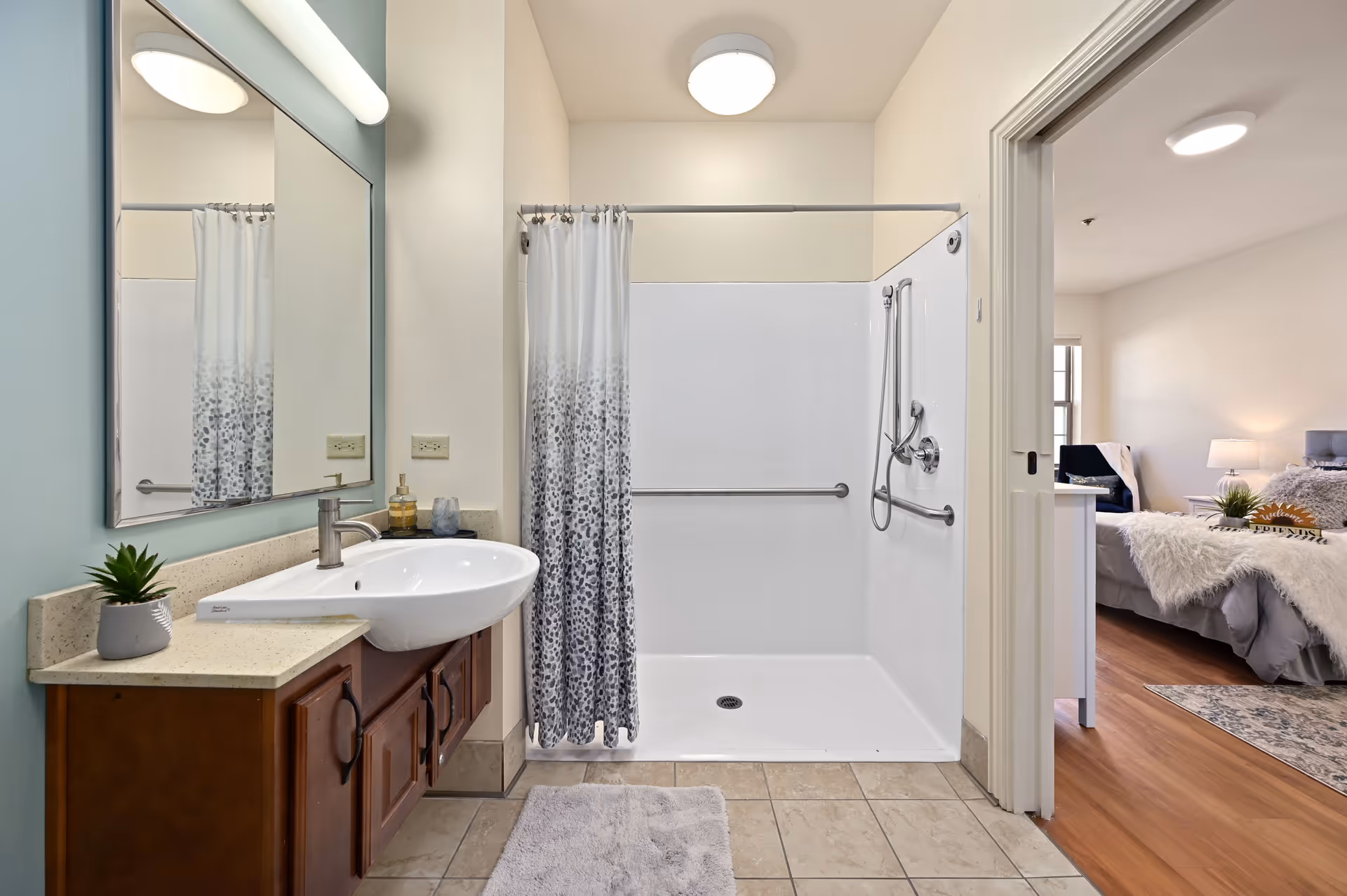 A bathroom with a large mirror above a sink with a wooden cabinet. There is a walk-in shower with a white and gray patterned curtain and grab bars. The bathroom floor is tiled, and a gray bath mat is placed in front of the sink. Through an open door, a bedroom with a bed, nightstand, lamp, and a rug on wooden flooring is visible.