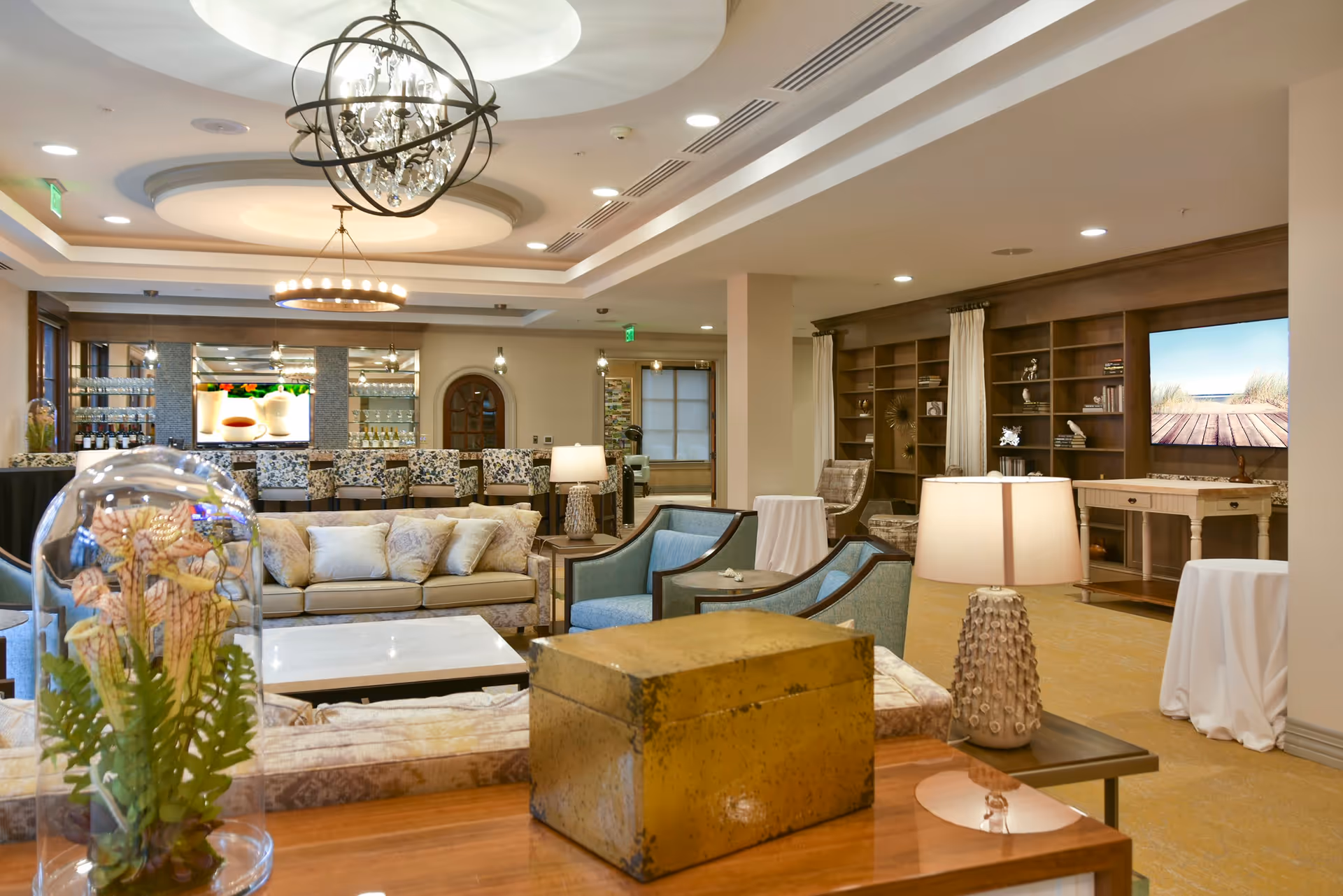 A spacious and elegantly decorated living room area in a senior living facility with comfortable sofas, armchairs, a coffee table, and decorative lamps. The room features a large built-in wooden bookshelf, a flat-screen TV displaying a beach scene, and a bar area with patterned bar stools and glass shelves. The ceiling has recessed lighting and two stylish chandeliers.
