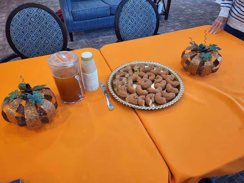 Orange-covered table in a communal room with a pitcher and stacked cups, a tray of crescent pastries, two decorative pumpkins, chairs in the background and a person's hand at the table edge.