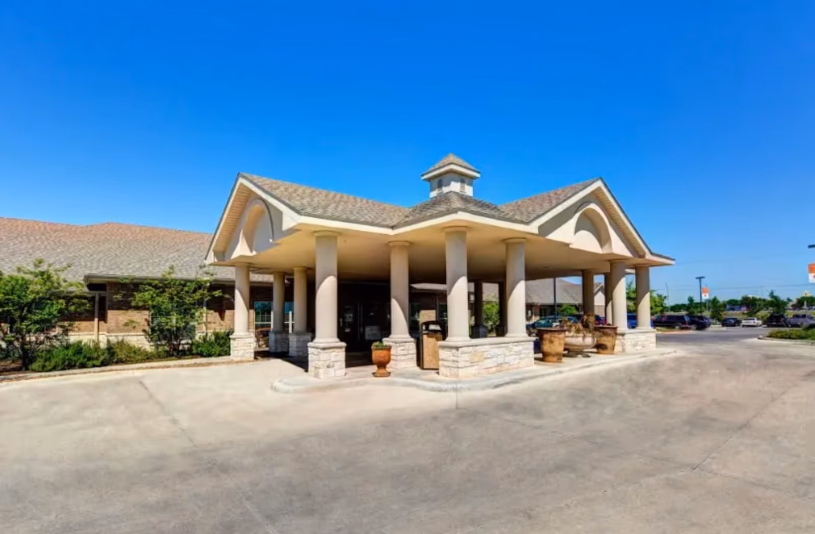 Exterior view of Hunters Pond Rehabilitation and Healthcare Center showing the main entrance with a covered driveway supported by large columns, surrounded by a parking lot and clear blue sky.