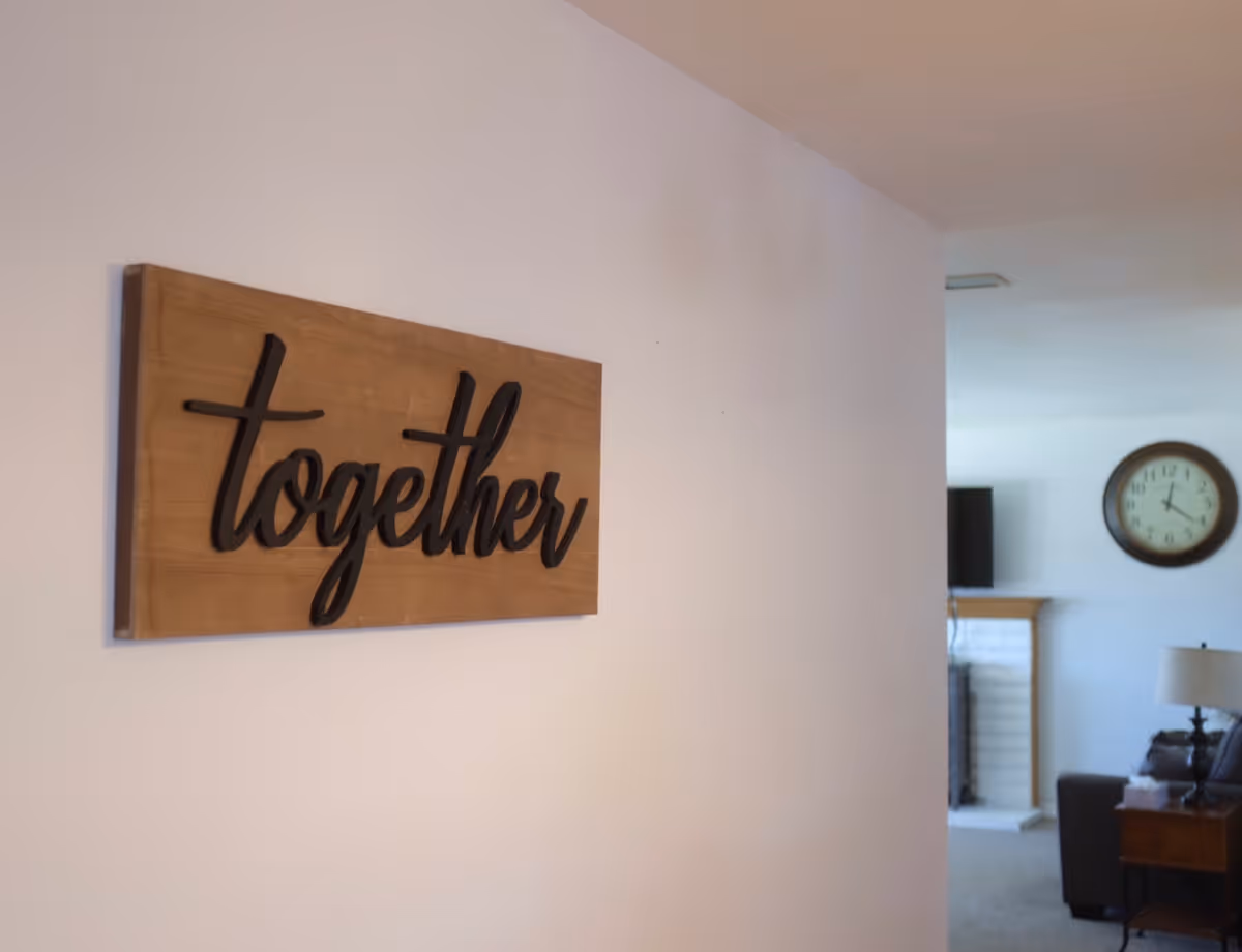 A wooden wall decoration with the word 'together' in black cursive letters mounted on a white wall. In the background, there is a living room area with a clock on the wall, a television above a fireplace, a lamp on a side table, and a couch.