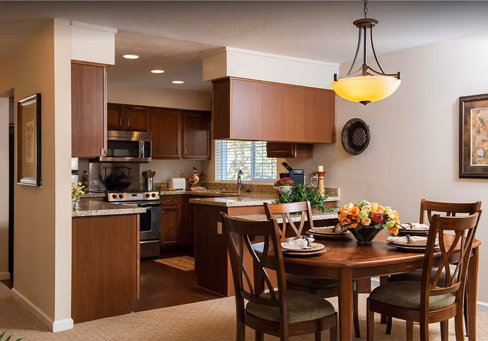 A cozy dining area with a wooden round table set for four, featuring a floral centerpiece. Adjacent to the dining area is a kitchen with wooden cabinets, granite countertops, a stainless steel stove, and a microwave. The kitchen has a window with blinds and recessed lighting.