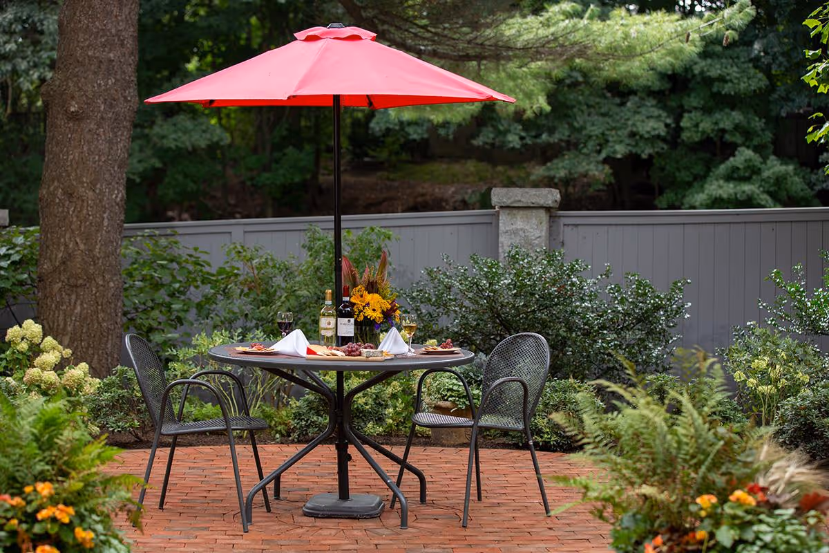 Outdoor patio area with a round metal table set for two, featuring wine bottles, glasses, napkins, and a flower arrangement under a red umbrella. The patio is surrounded by greenery and a gray fence.