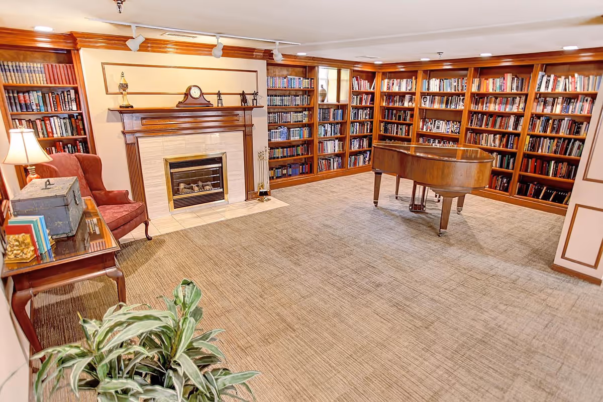 Cozy library-style common room with built-in bookshelves, a fireplace, a grand piano, an armchair, and a side table.