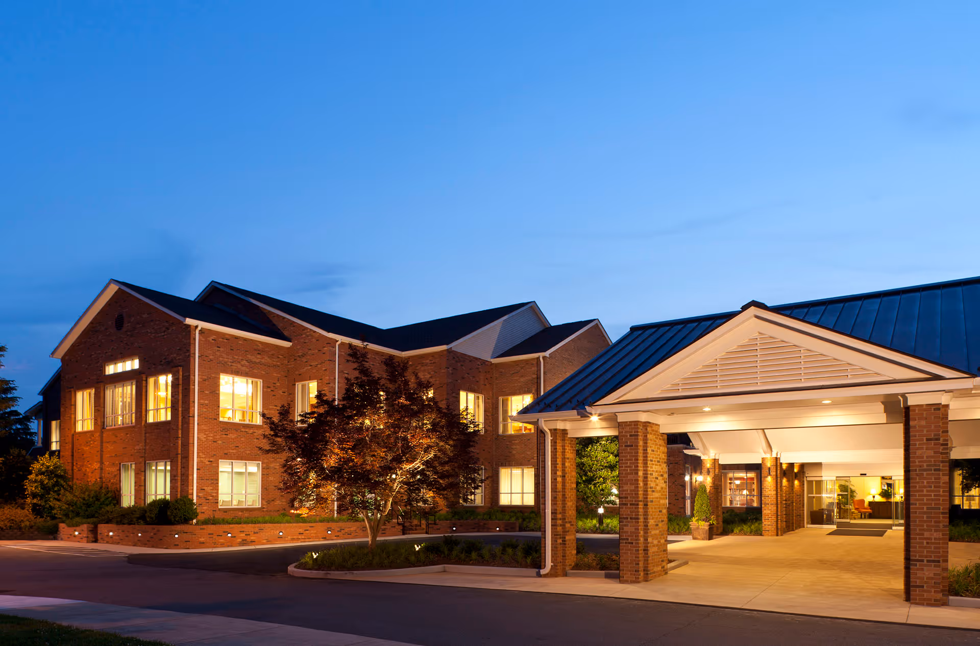 Brick senior living building at dusk with illuminated windows and a covered main entrance.