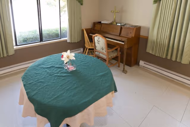 A small room with a round table covered with a green tablecloth and a beige undercloth, a vase with white and red flowers on the table, a wooden piano against the wall with a wooden chair in front of it, green curtains on the windows, and beige walls and floor.
