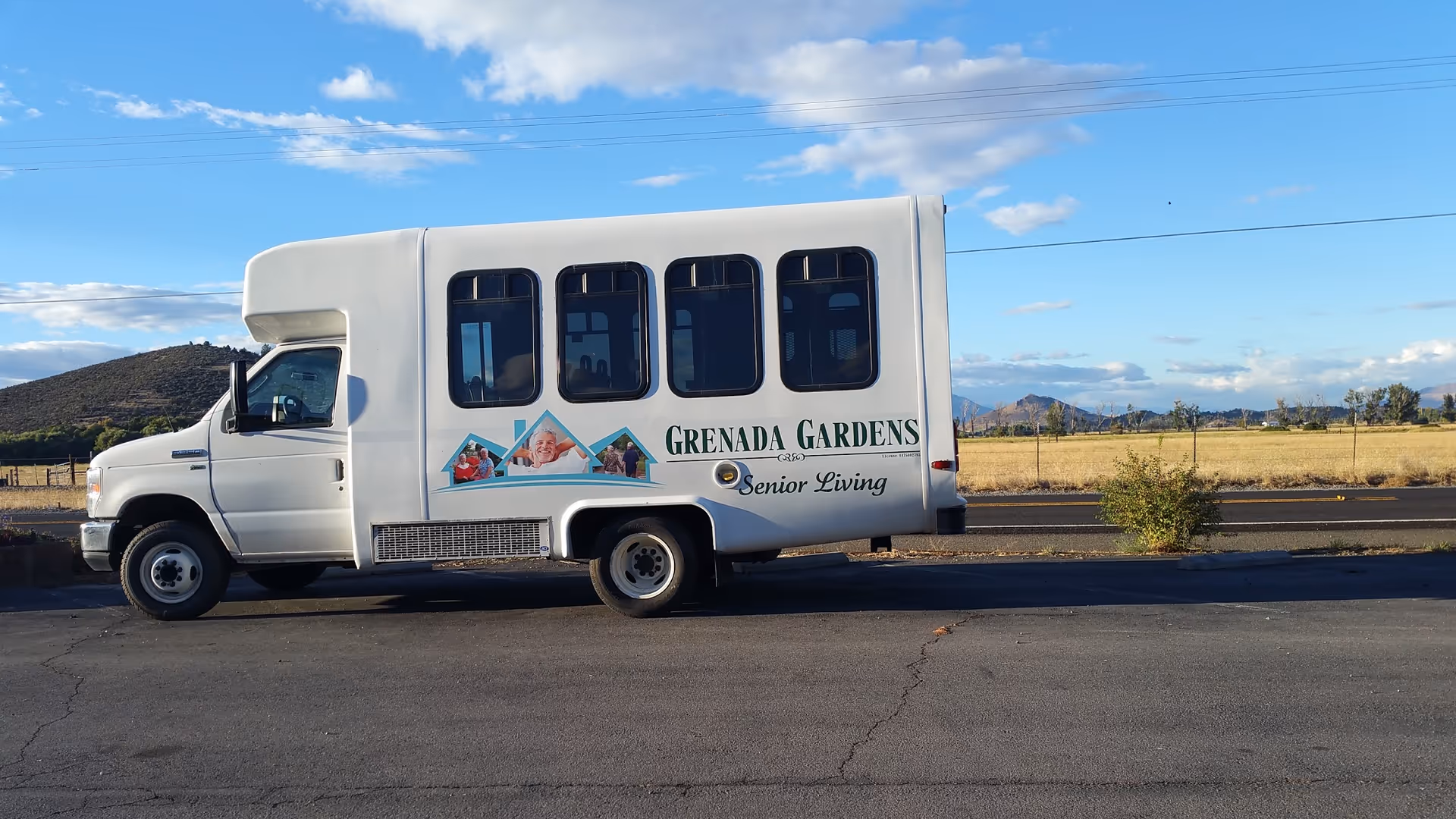 A white shuttle bus parked on a paved area with a scenic background of fields, hills, and a partly cloudy sky. The side of the bus displays the logo and text 'Grenada Gardens Senior Living' along with images of smiling seniors.