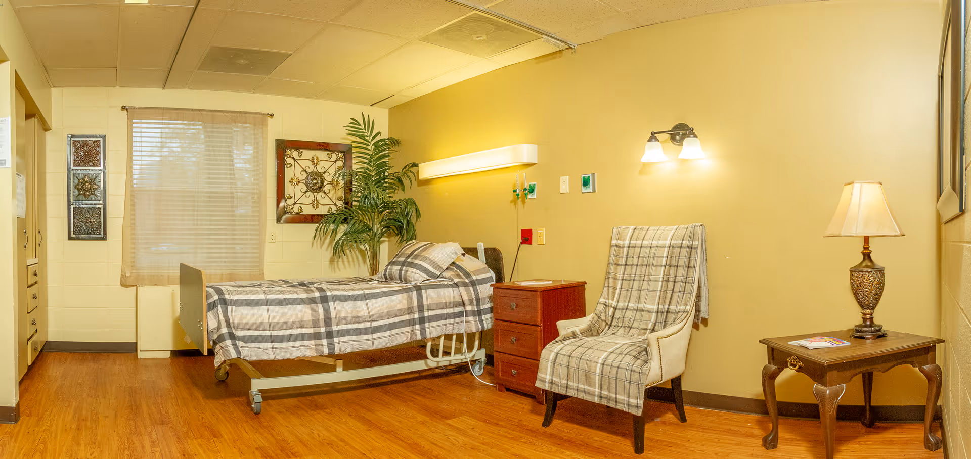 A well-lit nursing facility room with a single bed covered in a plaid blanket, a wooden nightstand with a lamp, a comfortable armchair draped with a matching plaid throw, a small wooden table with a decorative lamp and magazines, and a potted plant near the window with sheer curtains.
