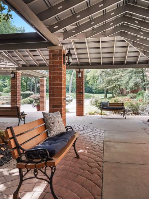 Covered outdoor seating area with wooden benches featuring cushions and pillows, supported by brick columns with hanging lanterns, overlooking a landscaped garden with trees and shrubs.