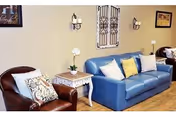 A cozy living room area with a blue couch adorned with multiple pillows and a brown leather armchair with a patterned cushion. Between them is a small white side table with a white orchid plant. The beige wall behind features decorative wall sconces and framed artwork.